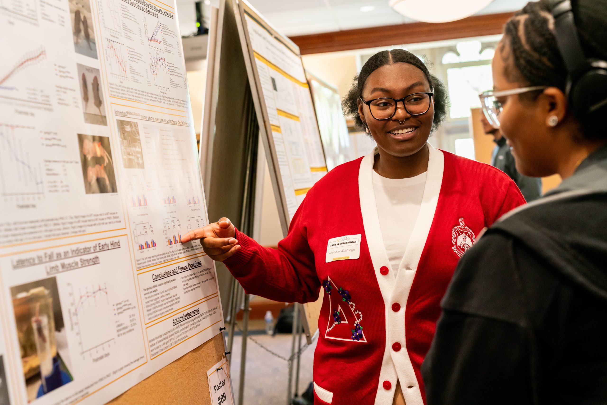 A student points to her research poster while describing her work to a second student.